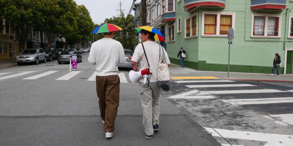 Two Boppn organizers walking a San Francisco street in umbrella hats, carrying a megaphone